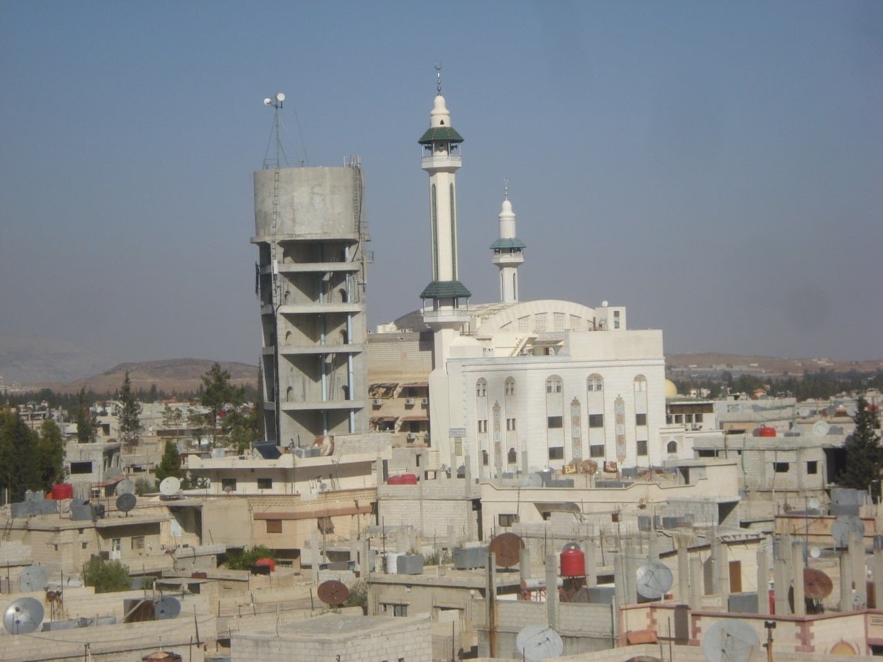 The rooftops of Khan Al-Sheikh Camp — water tanks, satellite dishes, and the mosque minarets above the hills of Rif Dimashq.